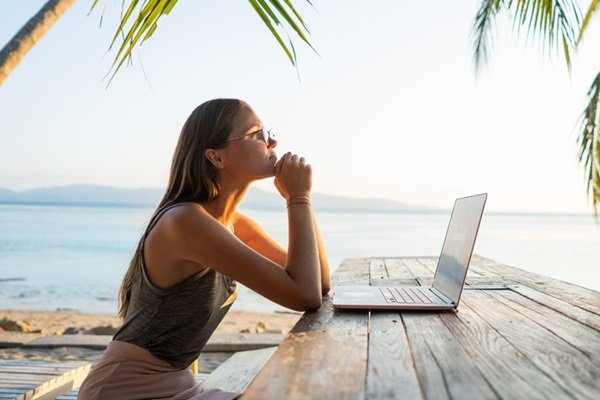 Woman relaxing with a laptop beside the beach, facing towards the text